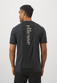 Schwarzes T-Shirt mit kurzen Ärmeln, das auf der Rückseite ein auffälliges graues "ellesse"-Logo vertikal zeigt. Es besteht aus einem glatten, leichten Stoff mit kontrastierenden Paneelen.