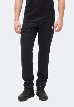 Férfi fekete kültéri nadrágot visel cipzáros oldalzsebbel és fekete túracipővel, világos, egyszínű háttér előtt áll.