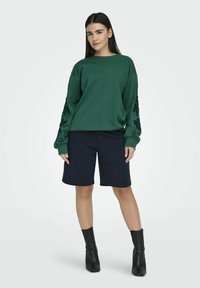 Πράσινο oversize φούτερ με κεντημένα λουλούδια στα μανίκια, συνδυασμένο με ναυτικές βερμούδες και μαύρα μποτάκια μέχρι τον αστράγαλο. Λείος υφασμάτινος ιστός.