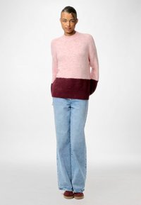 Ružičasta i burgundska pletena bluza u blokovima boja s dugim rukavima, u kombinaciji s svijetloplavim širokim trapericama i tamnocrvenim cipelama. Mekana tekstura.