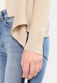 Beige ribbad cardigan med öppen front, med texturerad stickad tyg. Bärs över ljusblå denim med en kontrasterande marinblå och vit rem.