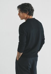 Černý dlouhý svetr s dlouhým rukávem a kulatým výstřihem. Hladká textura, přiléhavý střih, žebrované manžety a lem. Model stojí zády, ruce má v kapsách.