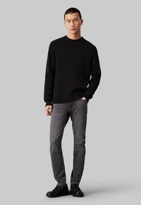 Maglione nero a coste con maniche lunghe e scollo a girocollo, abbinato a jeans grigi e scarpe in pelle nera, presentato su uno sfondo neutro.