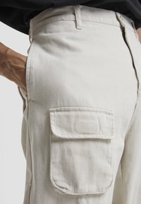Ljust beiga cargobyxor har ett slätt, texturerat tyg med sidofickor och en figursydd midja, och visar detaljerad sömnad och robusta sömmar.