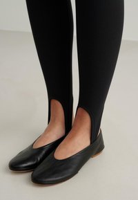 Svarta läderballetskor med rund tå och utskurna sidor, kombinerade med kroppsnära svarta leggings. Slät textur och minimal detaljering.