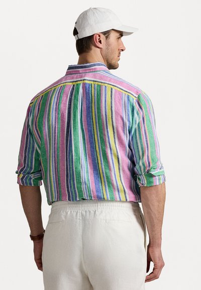 Chemise colonnée avec des rayures verticales multicolores en rose, vert, bleu et jaune. Manches retroussées et tissu léger. Casquette blanche portée.