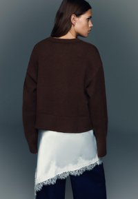 Pulover maro scurtat, cu textură moale și mâneci largi, purtat peste un articol vestimentar alb din satin cu dantelă la margine.