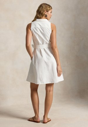 Kvinne står, iført en hvit ermeløs kjole med belte og krage, brune flate sandaler og svarte hoop-øredobber, mot nøytral bakgrunn.