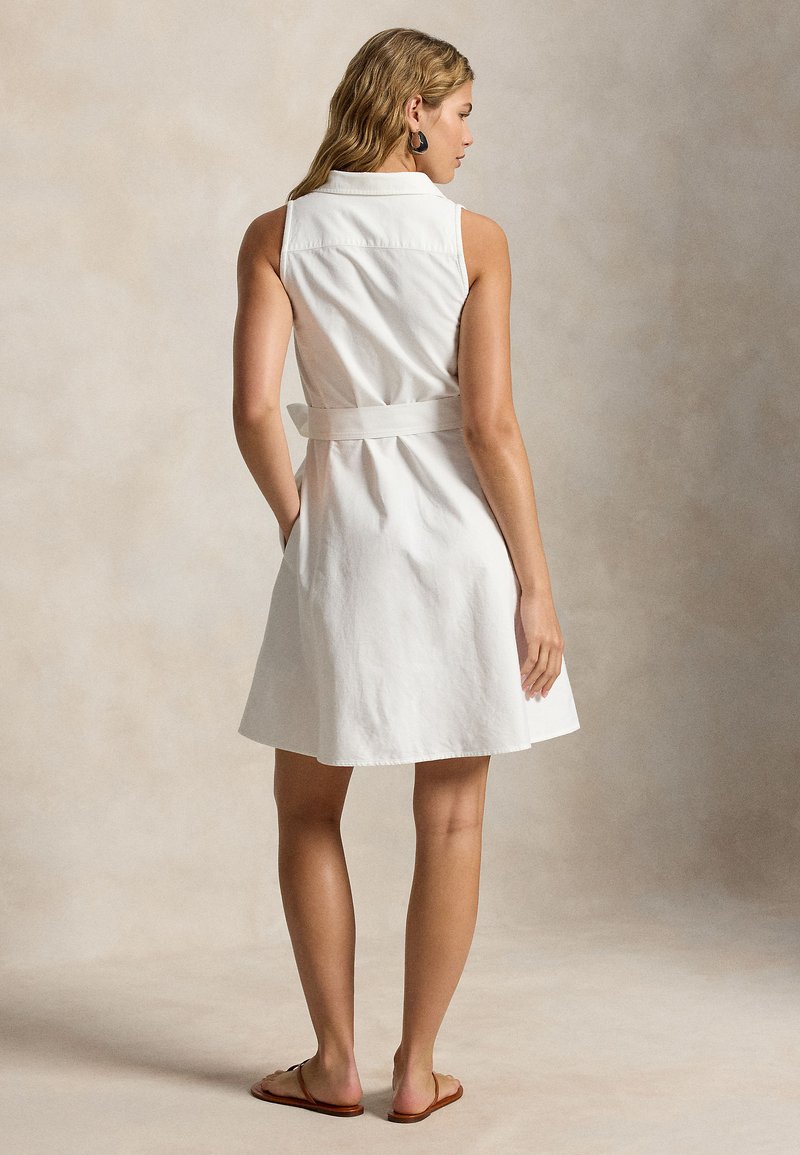 Femme debout, portant une robe blanche sans manches ceinturée avec col, des sandales plates marron et des boucles d'oreilles créoles noires, sur un fond neutre.