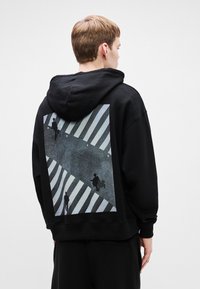 Persona vestido con una sudadera negra con un gráfico impreso de personas cruzando un paso peatonal con rayas en la parte posterior.