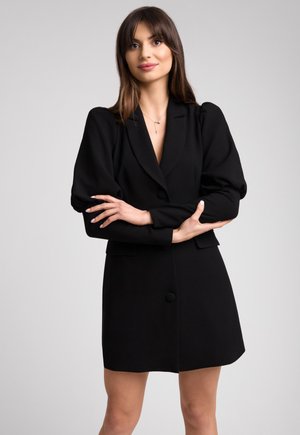 Kobieta z długimi brązowymi włosami, ubrana w czarną sukienkę-żakiet z bufiastymi rękawami, stojąca ze skrzyżowanymi ramionami na tle jasnej, jednolitej ściany.