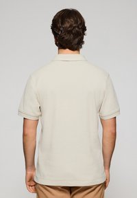 Polo shirt beige con colletto classico e maniche corte. Il tessuto appare liscio con una vestibilità rilassata, maniche orlate e una linea diritta.