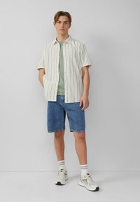 Kurzarm gestreiftes Hemd in Beige und Weiß, kombiniert mit einem grünen Rundhalsausschnitt-T-Shirt, blauen Jeansshorts und weißen Sneakers mit grauen Akzenten.