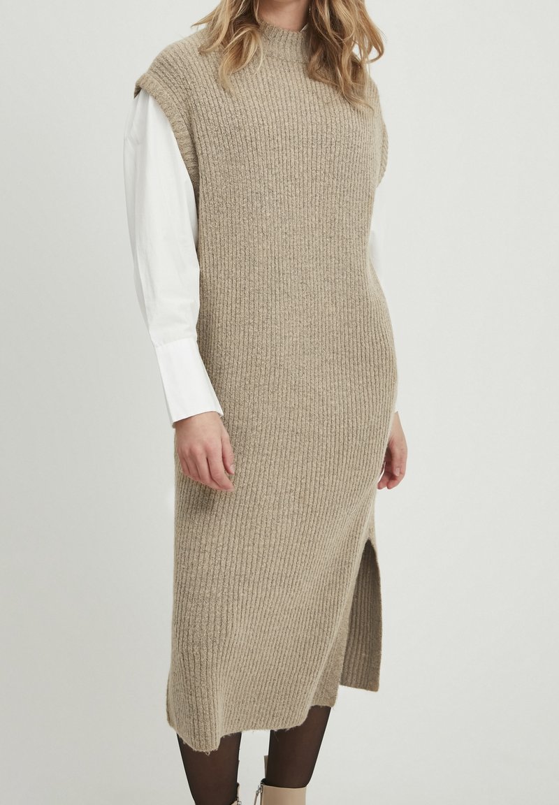 Femme portant une robe midi en maille côtelée beige sans manches par-dessus une chemise blanche à manches longues avec des collants noirs sur un fond uni.