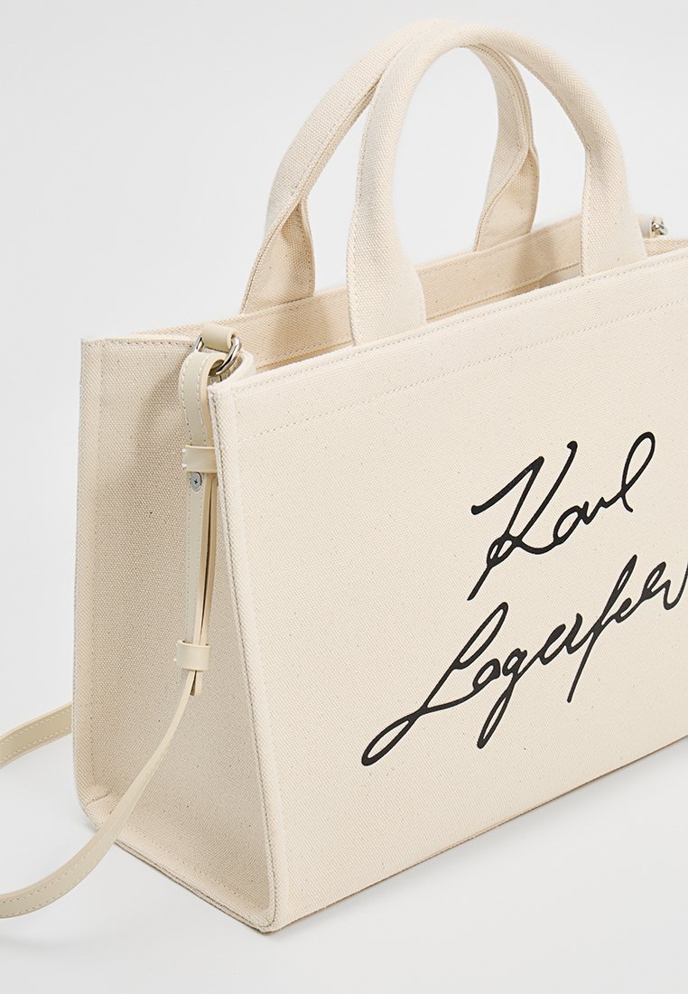 Sac fourre-tout en toile beige avec des poignées supérieures doubles et une bandoulière réglable. Présente une marque en cursive noire à l'avant. Texture simple.