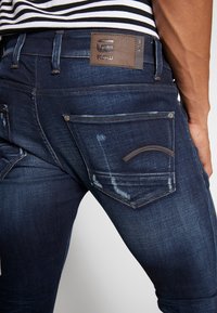 Dunkelblaue Denim-Jeans mit leichter Ausbleichung, ausgestattet mit einem Lederpatch, das "G-STAR RAW" beschriftet ist, sowie zwei Gesäßtaschen mit dekorativen Nähten.