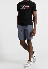 Schwarzes T-Shirt mit einem roten und weißen norwegischen Flaggenmuster, kombiniert mit grauen Shorts und weißen Turnschuhen mit schwarzen Streifen.