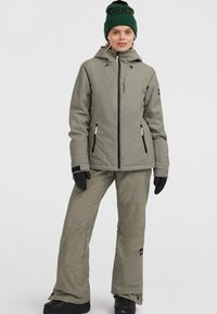 Grijze gestructureerde skijas met capuchon, gecombineerd met bijpassende skibroek; heeft zwarte ritsen en handschoenen, en een groene gebreide beanie met logo.