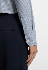 Lichtblauw overhemd met ruitjes, mouwen met knoopmanchetten, gecombineerd met donkerblauwe, gestructureerde broek. Belangrijke details zijn het patroon en het kleurcontrast.