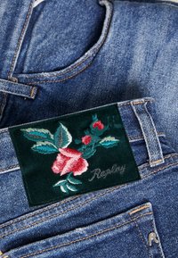 Farkut sinisessä pesussa sisältävät tummanvihreän samettitaskun, johon on brodeerattu vaaleanpunaisia ruusuja ja teksti "Replay". Vaurioituneet yksityiskohdat lisäävät tyyliä.