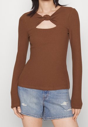 Frau trägt ein braunes, langärmliges, geripptes Oberteil mit Knoten-Detail und Ausschnitt an der Brust, kombiniert mit hellblauen, ausgewaschenen Denim-Shorts.