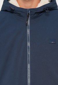 Giacca a zip blu navy con cappuccio, dotata di un morbido rivestimento in felpa beige e un piccolo patch con logo sul petto. Esterno in tessuto texturizzato.