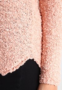 Gros plan d'une personne portant un pull rose texturé en maille en boucle avec des manches longues et un pantalon noir.