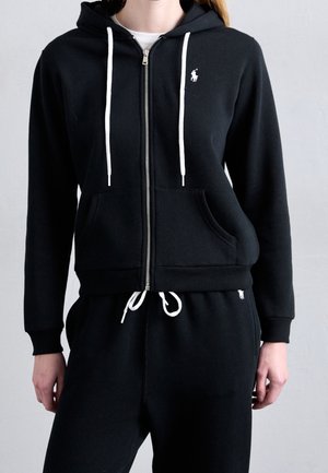 Woman wearing black zip-up hoodie with white drawstrings and matching black sweatpants with white drawstring ties, standing against a plain background.