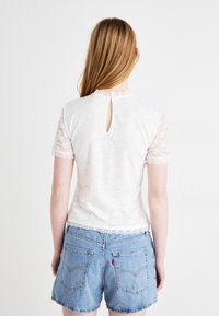 Hvit bluse i blonde med korte ermer, bølget halsutskjæring og spalte detaljer bak, kombinert med lyseblå denimshorts.