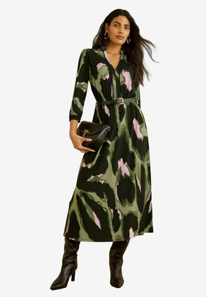 Kobieta nosząca sukienkę z długim rękawem w zielono-różowy abstrakcyjny wzór, czarny pasek, czarne kozaki na obcasie i trzymająca czarną torebkę.