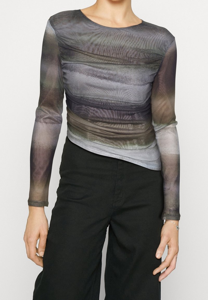 Personne portant un haut à manches longues semi-transparent avec des motifs abstraits gris et marron, associé à un pantalon noir taille haute et un bracelet.