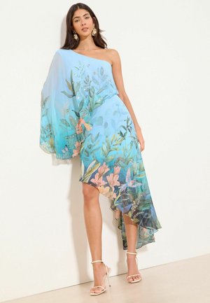 Vrouw draagt een blauwe jurk met één schouder met bloemen- en vogelprints, voorzien van een asymmetrische zoom en doorschijnende, vloeiende stof aan één mouw.