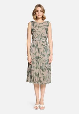 Frau steht und trägt ein ärmelloses beiges Kleid mit grünen Blattmustern, einem passenden Gürtel und weißen Sandalen vor einem schlichten hellen Hintergrund.