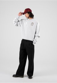 Felpa grigia con logo e numero 3, abbinata a pantaloni plissettati neri e un cappellino bordeaux. Presenta un fit rilassato e un design casual.