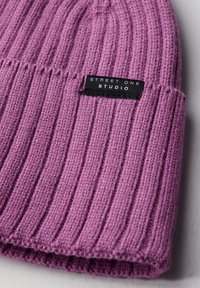 Berretto in maglia rosa con trama a costine. Presenta un'etichetta nera con testo bianco che recita "STREET ONE STUDIO" vicino al bordo inferiore.