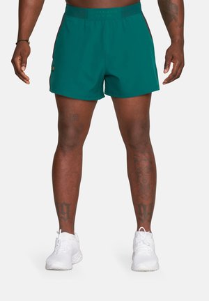 Mężczyzna w turkusowych krótkich spodenkach sportowych i białych trampkach, pokazujący tatuaże na nogach i obcięty tors na jednolitym tle.