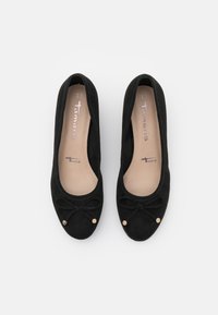 Černé semišové baleríny mají na špičce mašli a zlaté akcenty. Interiér je v světlé barvě s viditelnou značkovou etiketou.