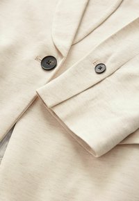 Beige bomuldsblazer med to sorte knapper, der viser en glat tekstur og struktureret revers. Nærbillede af knap og ærmedetalje.