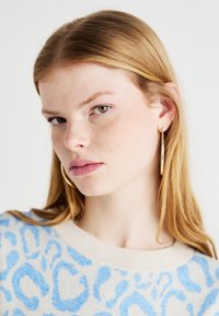 Πλεχτό πουλόβερ με μπεζ φόντο και μπλε λεοπάρ σχέδιο. Το μοντέλο φοράει χρυσά σκουλαρίκια κρίκους, έχει ίσια ξανθά μαλλιά και ανοιχτόχρωμο δέρμα με φακίδες.