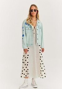 Kobieta ubrana w jasnoniebieską dżinsową kurtkę w niebieskie kropki, białą sukienkę midi w białe kropki, białe wysokie trampki i okrągłe okulary przeciwsłoneczne.