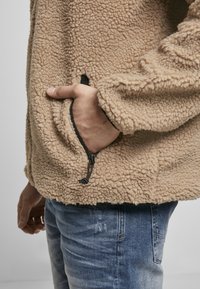 Beige fliisjakker, millel on tekstuuriga pind, küljetasku ja mustad aktsendid hemiku lähedal. Seljas on sinised teksad.