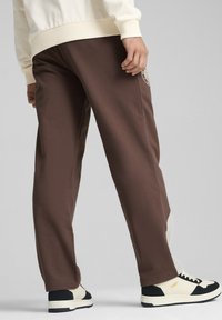 Braune Sweatpants mit lockerer Passform, mit einem Logo-Patch am linken Oberschenkel und kontrastierenden weißen Einsätzen. Kombiniert mit schwarzen und cremfarbenen Sneakers.