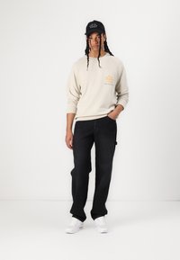 Beige Sweatshirt mit einem Sonnenmotiv und Text, schwarze Jeans und weiße Sneakers. Schwarze Kappe mit Text. Lange Haare in Zöpfen frisiert.