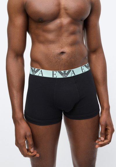 Αρσενικός κορμός που φορά μαύρα μπόξερ με ανοιχτό μπλε ελαστική ζώνη μέσης με μαύρο λογότυπο EA και εμβλημτικό αετό, στέκεται μπροστά σε απλό φόντο.
