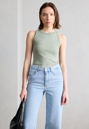 Νέα γυναίκα που φοράει αμάνικο τοπ σε απαλό πράσινο χρώμα και ανοιχτόχρωμα τζιν, κρατώντας μια μαύρη δερμάτινη τσάντα μπροστά σε απλό φωτεινό φόντο.