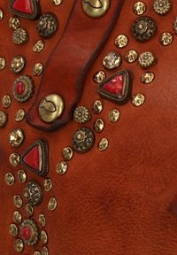 Superficie in pelle marrone decorata con borchie metalliche rotonde e triangolari. Presenta accenti rossi e texture varie. Notevole per le forme e le dimensioni diverse.