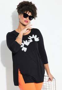 Tunic nera con applicazioni floreali bianche, maniche a tre quarti, scollatura rotonda e spacchi laterali. Abbinata a pantaloni arancioni.