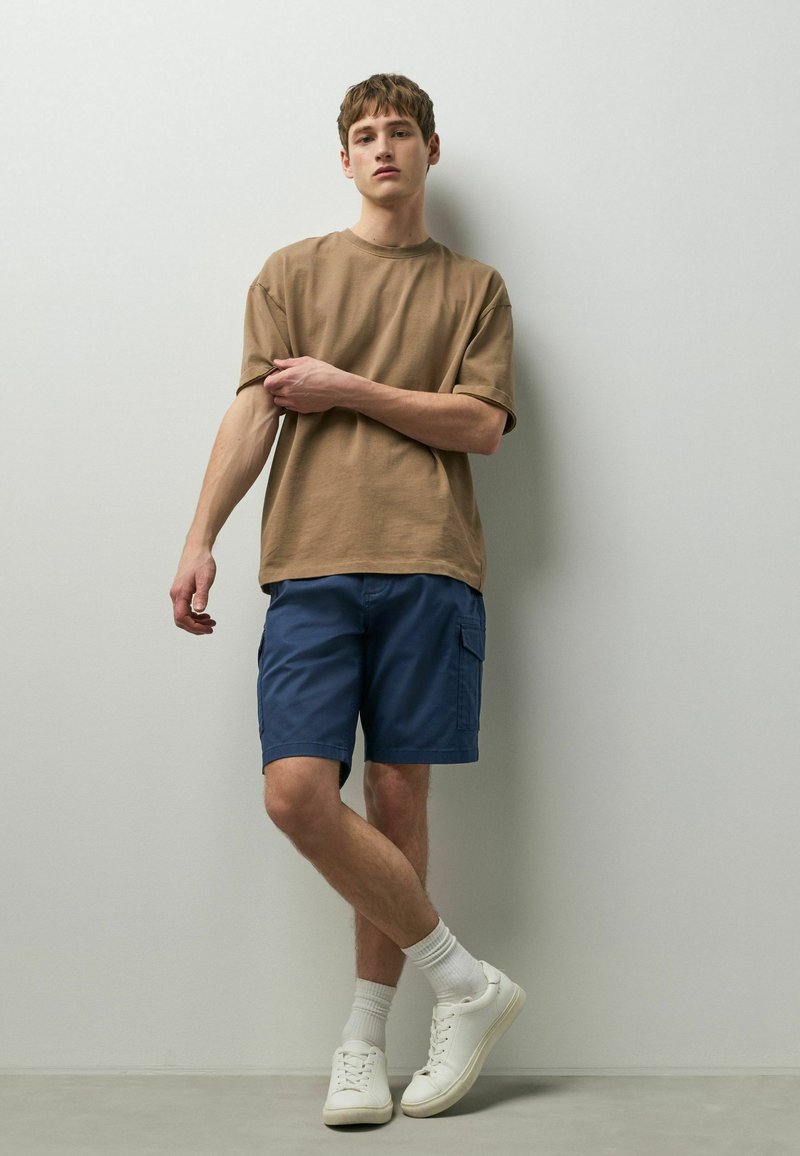 Jeune homme en t-shirt beige, short cargo bleu marine, chaussettes blanches et baskets blanches, debout devant un mur gris clair uni.