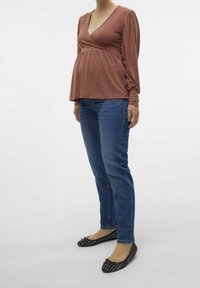 Těhotenský top z rzi v barvě s hlubokým V výstřihem a nařaseným empírovým pasem, spárovaný s modrými džínami a černými plochými botami.