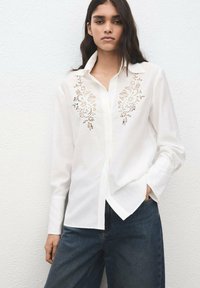 Camisa blanca de manga larga con detallado encaje en la parte frontal, diseño abotonado y corte relajado, combinada con jeans azul oscuro.